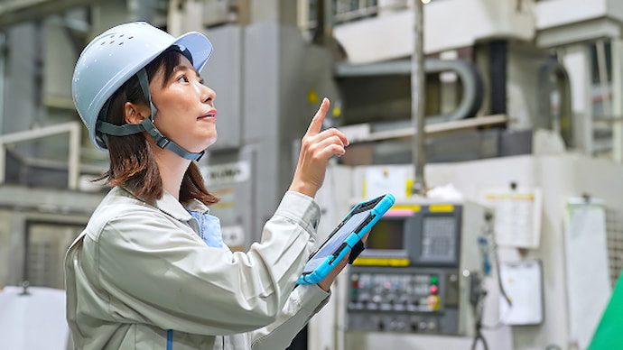 ヘルメット着用の女性が工場設備をタブレットで指さし確認している様子