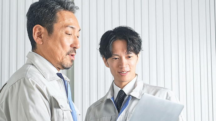 作業着姿の2人の男性がノートパソコンを見ながら話し合っている様子