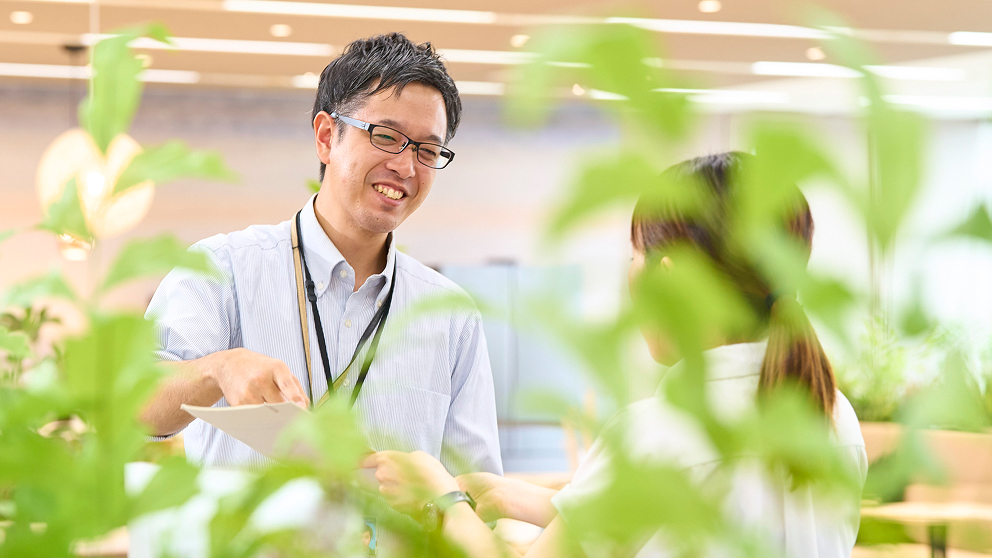 笑顔でオフィスで同僚と資料の確認をするD.N.さん
